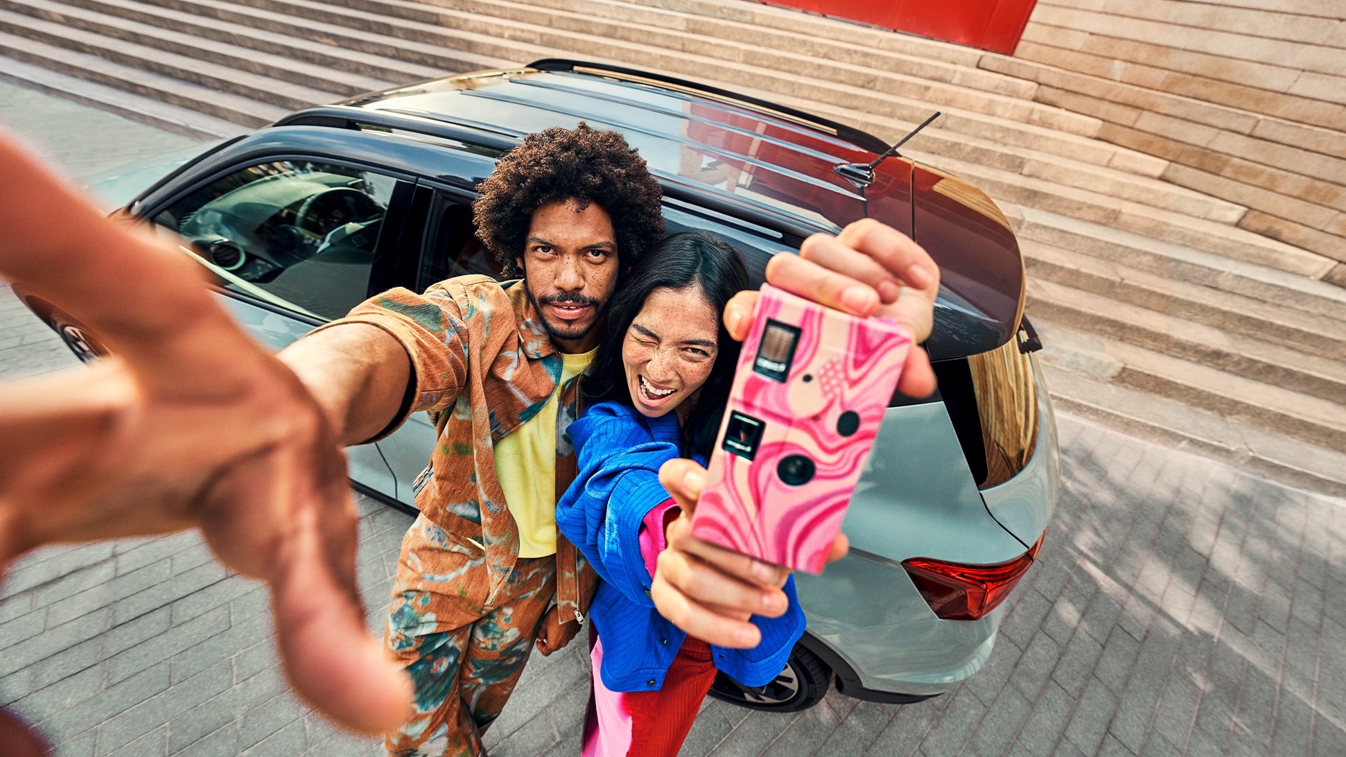 Two people taking a selfie in front of a SEAT Arona parked in an urban setting with modern steps and red wall in the background